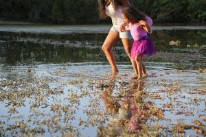Two girls in pink and white outfits wade barefoot through shallow golden-lit water with their reflections shimmering below.