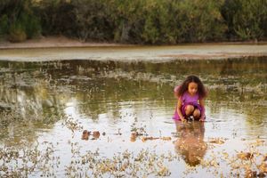 A young girl in a purple outfit crouches in shallow water, curiously exploring the pond's surface surrounded by green shrubs.
