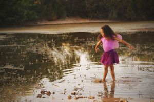 A girl in a pink top and purple tutu skirt walks through shallow pond water while sunlight reflects across the surface.