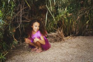 A smiling young girl with long curly hair in a pink top and purple skirt crouches on gravel beneath green leafy plants.