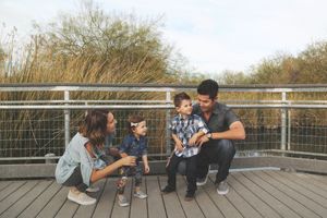 Parents crouch on a wooden boardwalk with their two young children, smiling together in front of tall golden reeds.