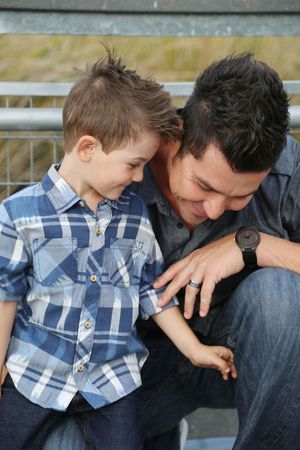 A smiling father in a gray shirt and watch leans down to hug his young son, who wears a blue plaid button-up shirt.