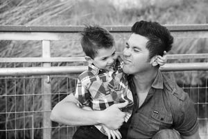 A black and white photo of a smiling father holding his young son in a plaid shirt, with a metal railing behind them.