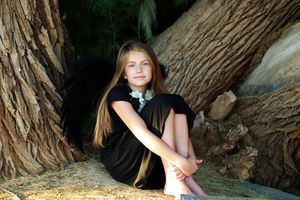 A long-haired girl in a black dress sits with her arms resting on her knees among the large, textured roots of a tree.