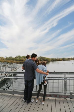 A family of four embraces on a wooden boardwalk, looking out over a calm lake under a wide blue sky with wispy clouds.