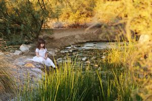 A young girl in a white dress sits on rocks beside a small stream, surrounded by tall green grass in warm golden light.