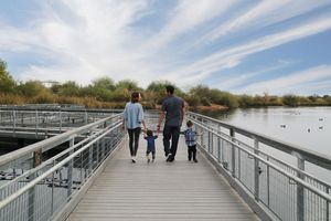 A mother and father walk hand in hand with their two young sons along a wooden boardwalk beside a peaceful lake.