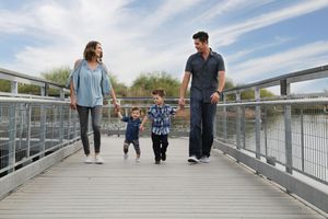 A smiling family of four, with two young sons in the middle, walks hand in hand along a lakeside boardwalk with railings.