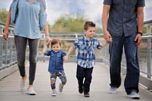 A toddler in a denim shirt and a boy in a plaid shirt hold their parents' hands while walking along a lakeside boardwalk.