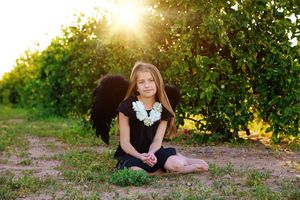 Girl in a black dress and black angel wings sits on a grassy path in an orchard with golden sunlight behind her.