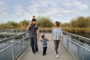 Family of four walks hand in hand along a wooden boardwalk by the water, with a child riding on the father's shoulders.