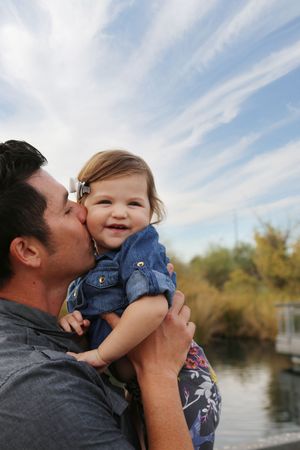 Smiling baby in a denim outfit is held and kissed on the cheek by her father near a lake under wispy clouds.