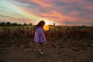 Little girl in a lavender dress twirls in a field of tall grass beneath a colorful pink and orange sunset sky.