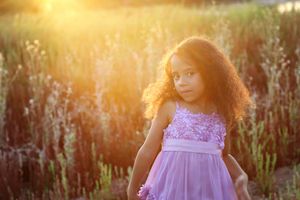Curly-haired girl in a purple lace dress stands in a golden field as warm sunset light glows behind her.