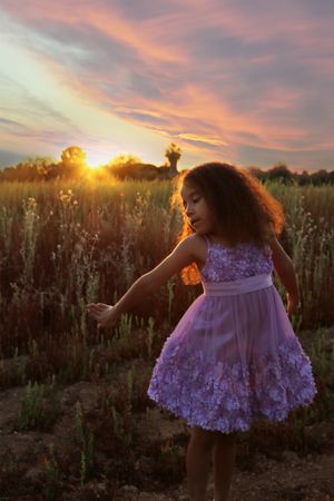 Girl with curly hair in a lilac floral dress dances in a meadow at sunset with the sun glowing on the horizon.