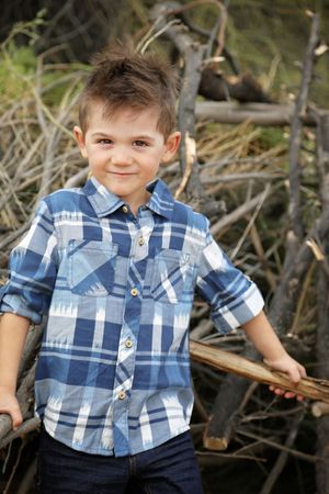 Smiling young boy in a blue plaid shirt and jeans stands in front of a pile of dry branches outdoors.
