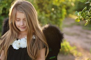 Girl with long hair wears black angel wings and a white flower necklace, looking down in a sunlit green orchard.