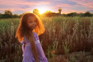 Smiling girl with curly hair in a purple dress stands in a field of tall grass as the sun sets in the background.