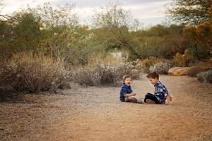 Wide view of two small children sitting on a dirt path surrounded by desert brush and trees.