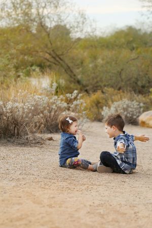 Two young children sitting on a sandy desert trail, laughing and playing together near shrubs.