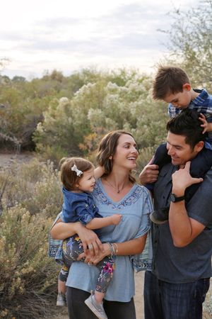 Smiling family of four outdoors, with a mother holding a baby girl and a father carrying a boy on his shoulders.