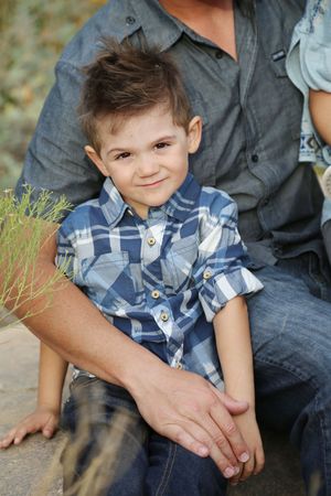 Smiling little boy in a blue plaid shirt sitting on his father's lap outdoors.