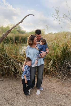 Family of four posing by tall grasses and a pond, with parents holding a baby and a boy standing nearby.
