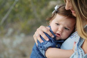 Mother tenderly hugging a baby girl in a denim outfit and white bow outdoors.