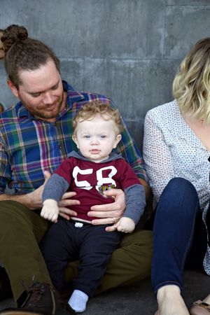 A smiling bearded man with a hair bun holds a curly-haired baby in a maroon shirt while a woman sits beside them.