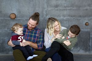 Smiling family of four sits against a gray concrete wall as parents hold and hug their baby and young son.