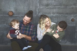 A happy family of four laughs and hugs on a concrete ledge, with dad in a plaid shirt and mom holding their boys.
