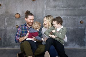 A smiling family of four sits together against a gray concrete wall, with parents cuddling their toddler and young son.