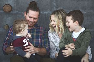 A laughing father and mother snuggle their toddler and young boy in a joyful family portrait against a concrete wall.