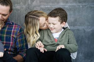 A mother kisses her smiling young son on the cheek while the family sits close together in front of a concrete wall.