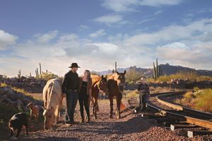Cowboy, woman and child stand with horses and dogs near railroad tracks in a desert landscape with cactus and mountains.
