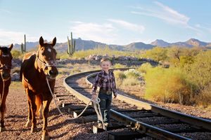 Young boy in a plaid shirt leads a brown horse beside curved railroad tracks in a sunny desert with saguaro cactus.