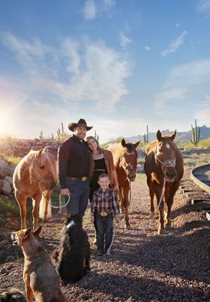 Cowboy, woman and young boy standing with three horses and two dogs on a desert path at sunset with cacti behind.