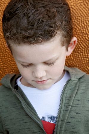 A close-up portrait of a young boy with curly brown hair looking down, wearing a green zip hoodie against a copper background.