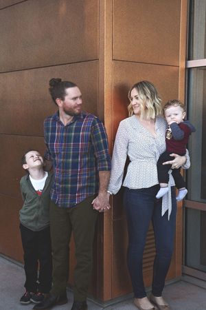 A family stands holding hands outside a modern building as a young boy looks up laughing, with parents and toddler smiling.