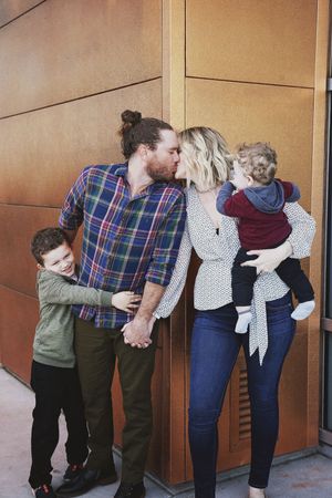 A father kisses a mother holding their toddler while their young son hugs them, standing beside a bronze-paneled building.