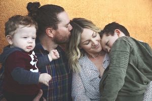 A close family portrait shows a bearded father kissing a smiling mother as their toddler and young son cuddle against a golden wall.