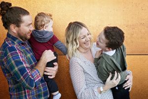 A father lifts their toddler as a laughing mother hugs their young son in a cheerful family photo against a golden wall.