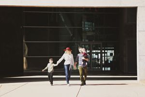 A family walks hand in hand toward a camera outside a modern building, with a mother in a red hat carrying their toddler.