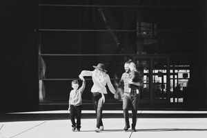 Black and white photo of a family of four walking hand in hand outside a modern glass building, with a mother in a wide-brim hat.