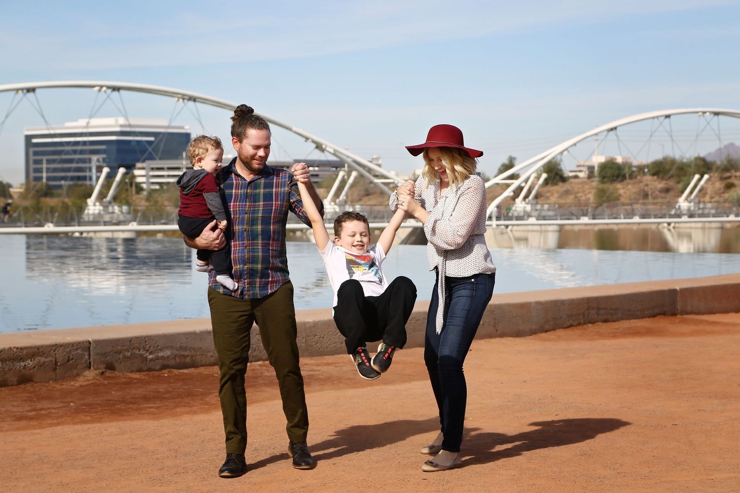 A family plays outdoors as parents swing a laughing boy by his arms while holding a baby near a modern arched bridge and reflecting pool.
