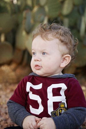 Curly-haired toddler in a maroon number 58 shirt with gray sleeves sits in front of prickly pear cactus, looking off to the side.