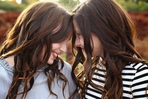 Two young women with long wavy brown hair touch foreheads and smile warmly in a tender outdoor portrait with a soft blurred background.