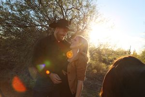A man in a cowboy hat and a blonde woman share a close moment outdoors among desert trees with golden sunset light and lens flare.