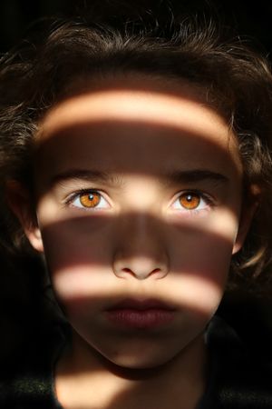Close-up portrait of a child with striking amber eyes as stripes of sunlight and shadow fall across the face against a dark background.