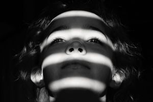 Black and white portrait of a child looking upward as striped light and shadow patterns cross the face in a dramatic artistic composition.